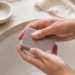 Mãos com espuma a segurar o sabonete de argila roxa Tradição Natural junto ao lavatório